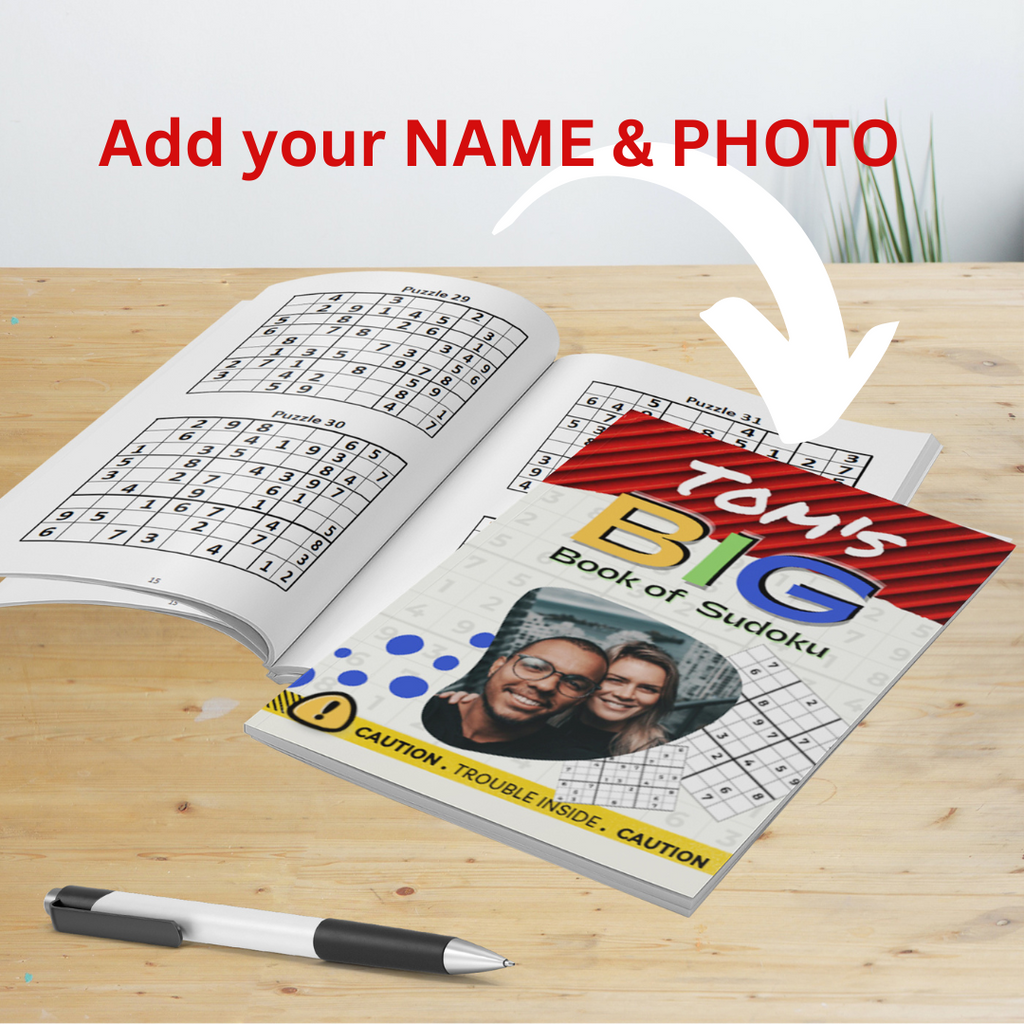 An informational image on a wooden table shows an open Sudoku puzzle book to the left with an arrow pointing to a closed, personalized version to the right. Red text above says, 'Add your NAME & PHOTO'. The cover of the closed book reads 'TOM'S BIG Book of Sudoku' and features a custom photo. A pen is in the foreground.