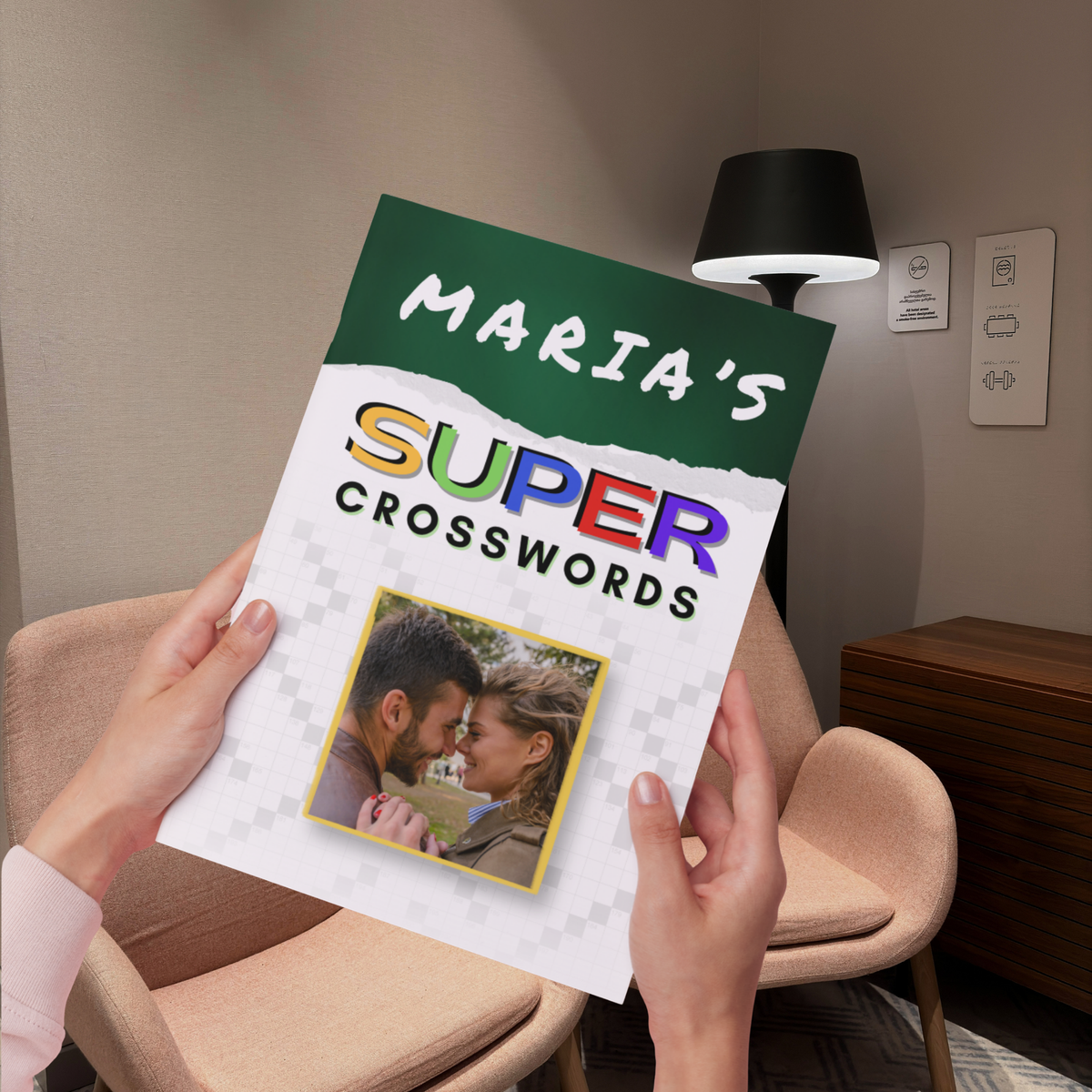 A person holds a printed book or magazine titled "Maria's Super Crosswords" in a cozy indoor room with beige chairs and a lamp in the background. The cover is white and green with colorful lettering and a small photo of a couple.