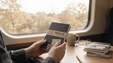 Man opens a book called 'Brad's Pocket Puzzeles' revealing the interior pages with crossword clues and crossword puzzle. 