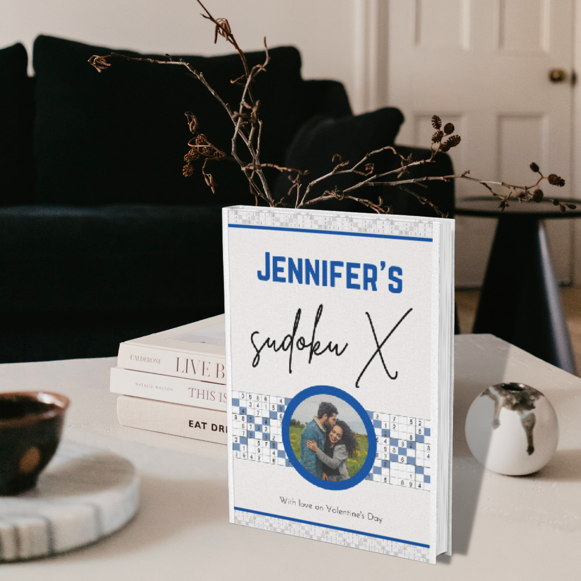 Personalized Sudoku X gift book in blue with a custom name and message on the cover, displayed on a table with decorative accents