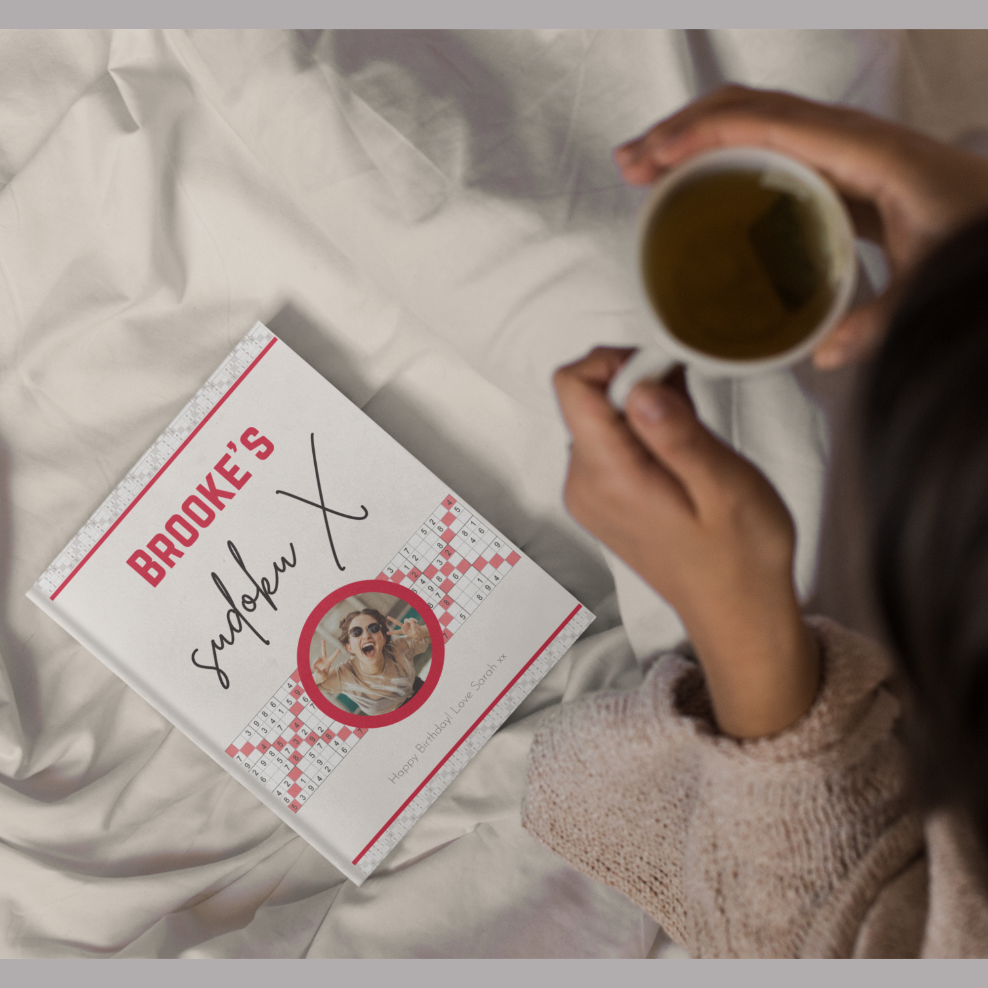 A lifestyle photo of a woman holding a mug; beside her on the bed is a red-themed puzzle book titled 'Brooke's Sudoku X'