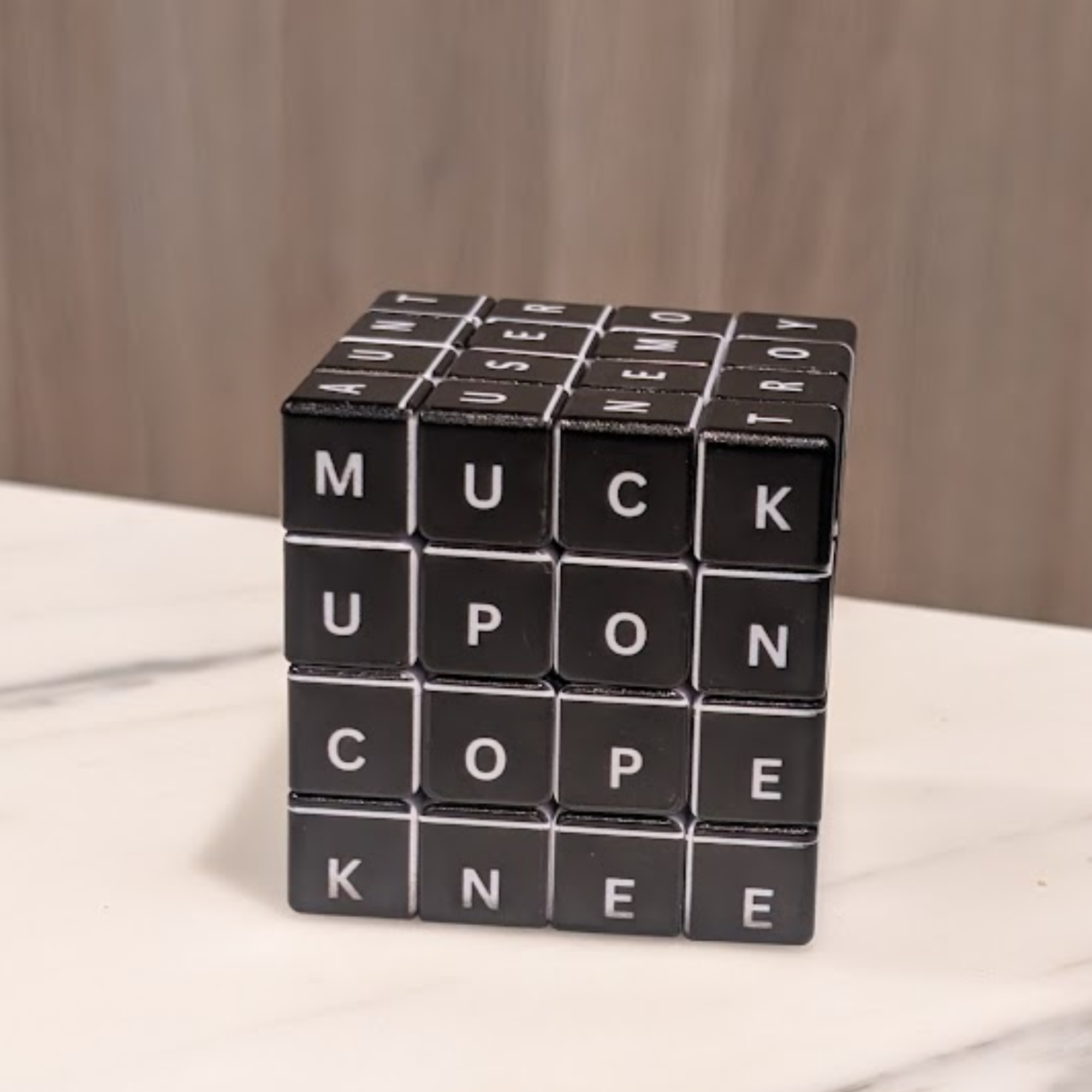A close-up of the 4x4x4 Witty Word Cube sitting on a white table. The image shows several sides of the cube with various letters aligned to form words