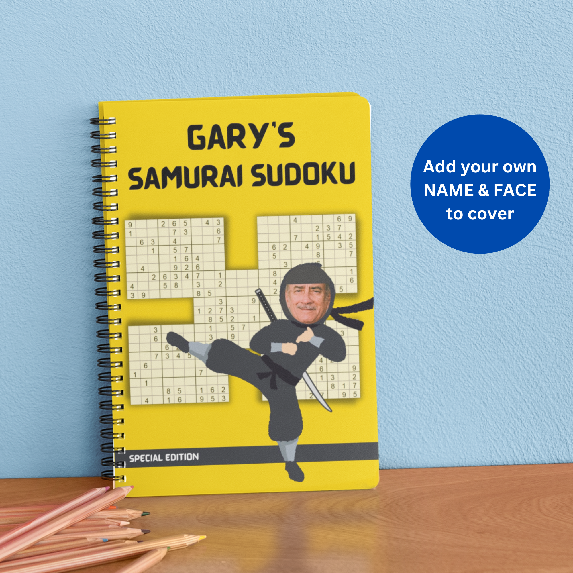 A wire coil-bound book with a bright yellow cover titled 'GARY'S SAMURAI SUDOKU,' showing an samuari sudoku puzzle grid and a ninja illustration with a man’s face; next to it is a blue circle graphic that says, 'Add your own NAME & FACE to cover.'