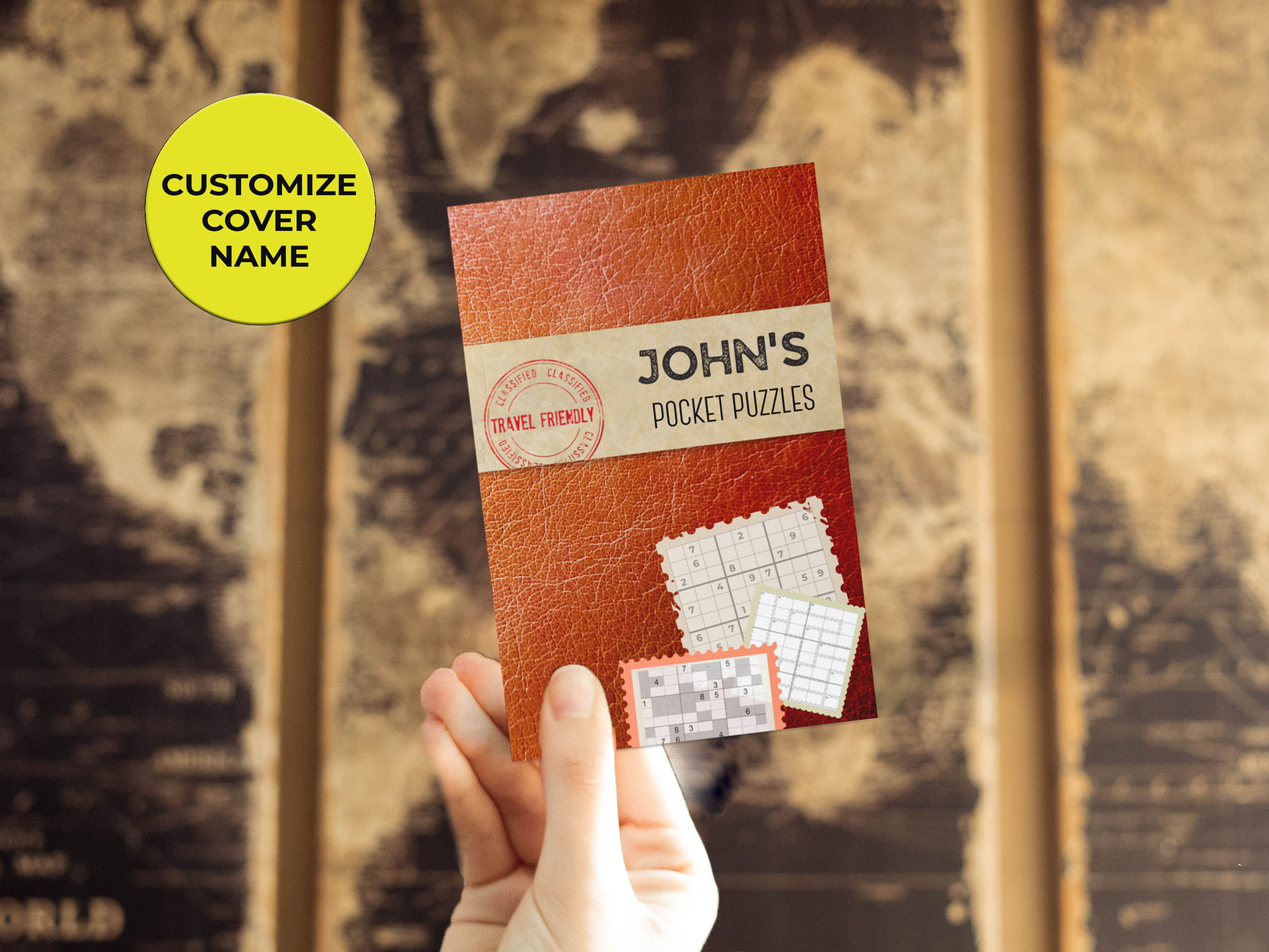 A hand holds a pocket-sized puzzle book titled 'John's Pocket Puzzles' in front of a world map. The cover has a leather texture, a 'Travel Friendly' stamp, and a yellow sticker that says 'Customize Cover Name'.