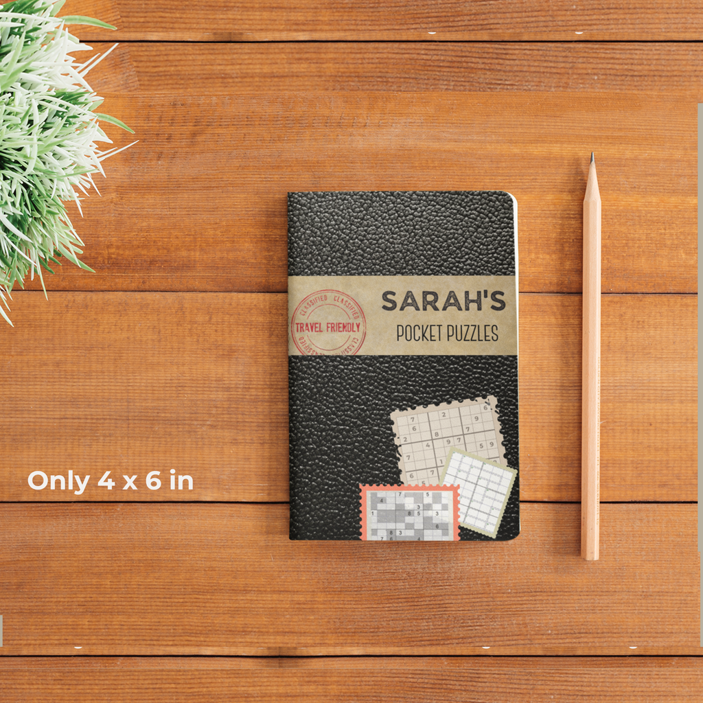 A small 4x6 inch black puzzle book titled 'Sarah’s Pocket Puzzles' sits on a wooden table. A pencil and a small green plant are nearby to show the compact scale. A stamp on the cover reads 'Travel Friendly'.