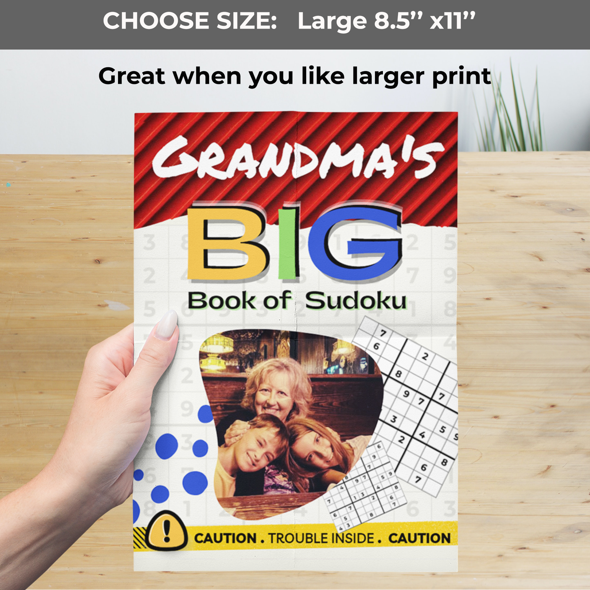 A hand holds a large Sudoku book over a wooden surface. Top text reads 'CHOOSE SIZE: Large 8.5” x 11”' and 'Great when you like larger print.' The cover features a photo of a grandmother with her family.