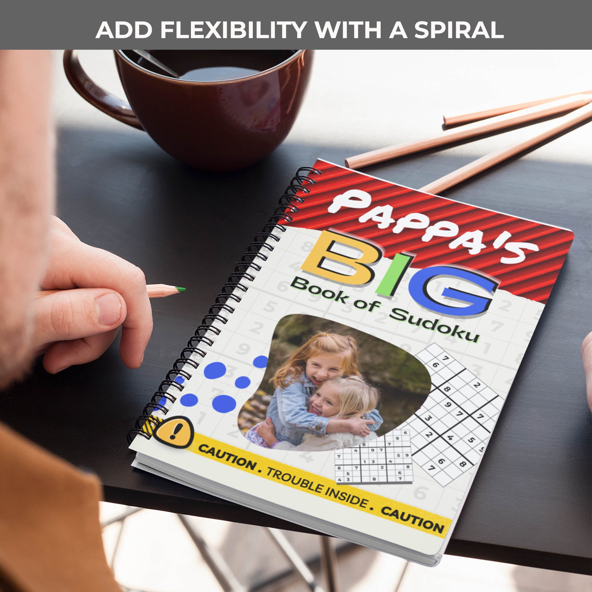 A lifestyle photo of a Sudoku book titled 'Papa's Big Book of Sudoku.' The image is angled to highlight the metal spiral binding along the spine, shown resting on a dark tabletop.