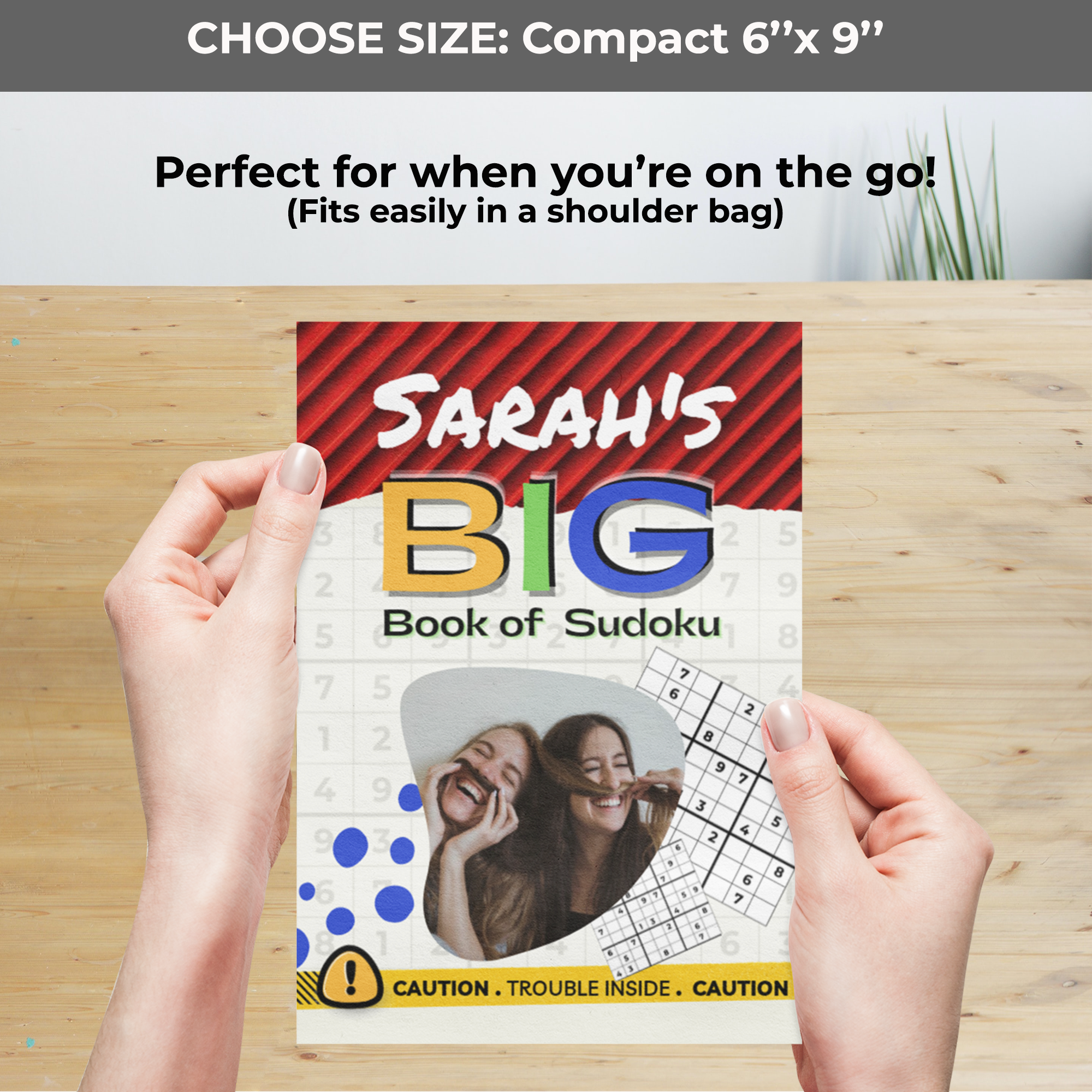Two hands hold a compact Sudoku book over a wooden surface. Top text reads 'CHOOSE SIZE: Compact 6” x 9”' and 'Perfect for when you’re on the go!' The cover shows two women laughing.